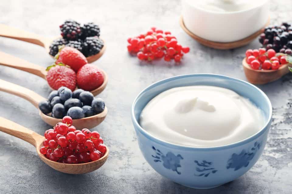 Naturjoghurt in einer Schüssel als Basis, verschiedene rote Früchte in Löffeln zur individuellen Ergänzung